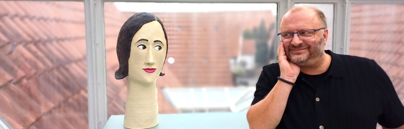 Ein Frauenkopf aus Pappmaché blinzelt zu einem neben ihm sitzenden Mann, der mit gleichem Gesichtsausdruck zurückschaut