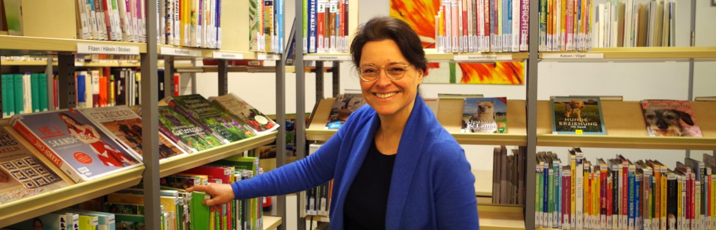 Gezeigt wird eine Mitarbeiterin, an einem Bücherregal stehend und in die Kamera lächelnd