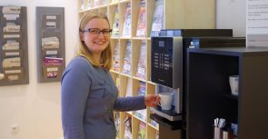 Gezeigt wird eine Dame an der Kaffeemaschine stehend