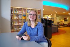 Eine junge Frau sitzt mit einem Tablet in der Hand im Lesedafé der Bibliothek Rheda und lächelt in die Kamera