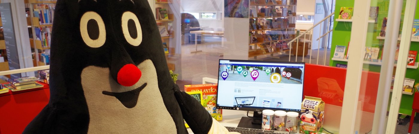 Ein großer Maulwurf als Stofftier sitzt an einem Schreibtisch mit PC