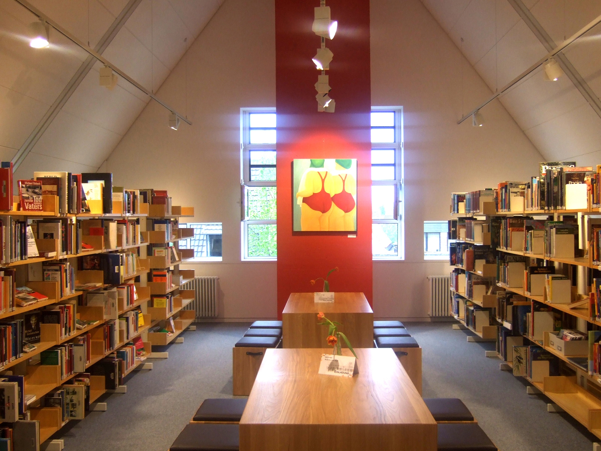 An einem rot angestrichenen Kamin im Galeriegeschoß der Bibliothek Wiedenbrück hängt ein ausgestelltes Bild der Artothek, rechts und links sind Bücherregale zu sehen