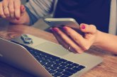 Gezeigt wird ein Laptop und Hände, die ein Smartphone in der Hand halten
