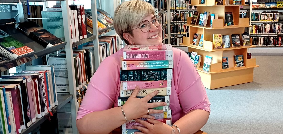 Eine Mitarbeiterin sitzt an einem Tisch und umarmt einen vor ihr liegenden Stapel Bücher