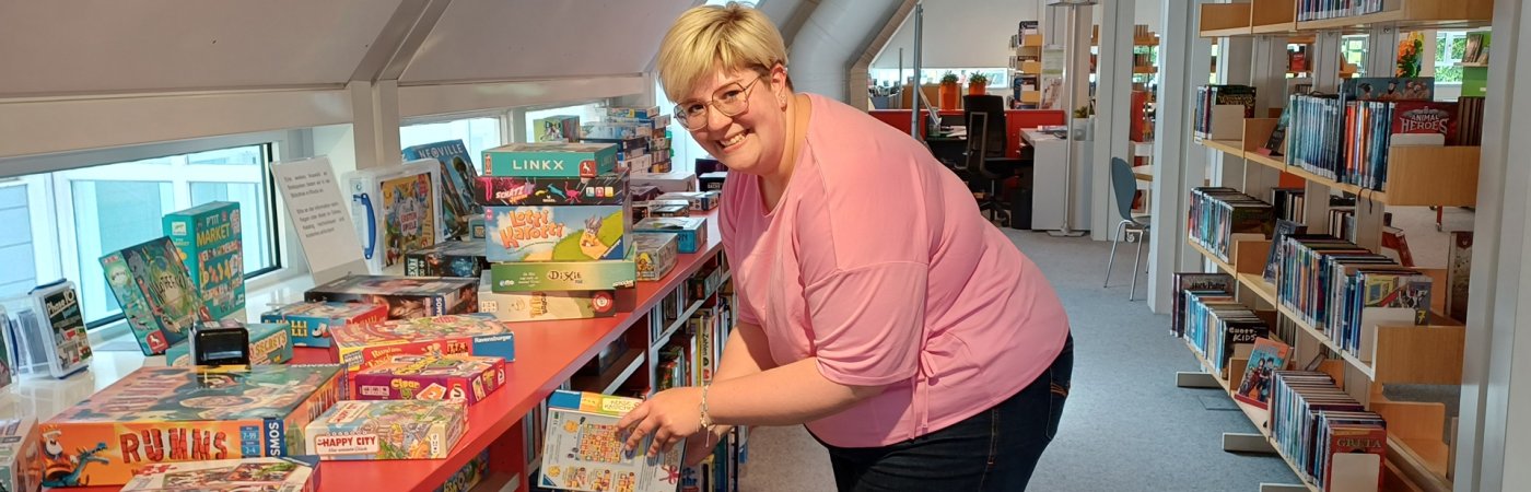 Eine Mitarbeiterin steht leicht gebeugt vor dem Brettspieleregal