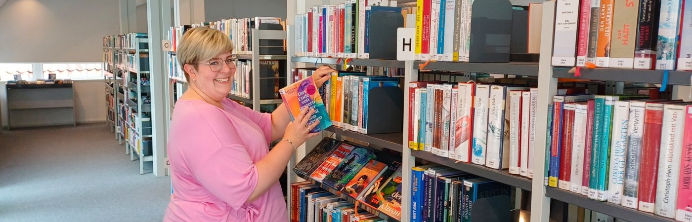 Eine Mitarbeiterin steht an einem Bücherregal und lächelt in die Kamera