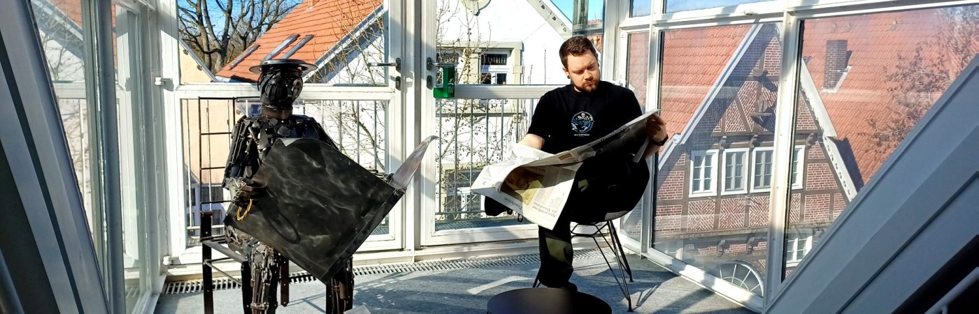 Gezeigt wird ein im Glaserker der Bibliothek in Wiedenbrück sitzender junger Mann, der Zeitung liest, daneben eine Metallfigur, die einen Zeitungsleser darstellt