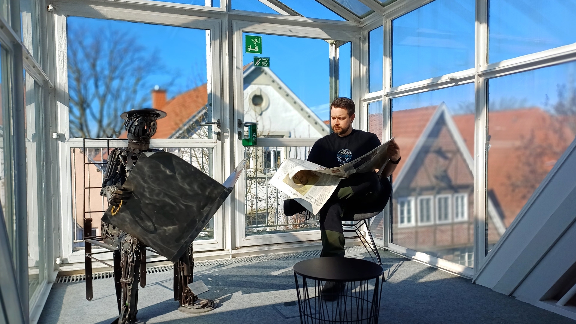 Gezeigt wird ein im Glaserker der Bibliothek in Wiedenbrück sitzender junger Mann, der Zeitung liest, daneben eine Metallfigur, die einen Zeitungsleser darstellt