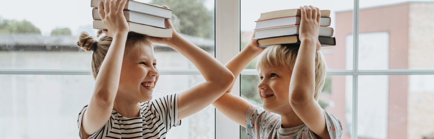 Zwei Kinder stehen sich einander anlachend gegenüber und halten sich Bücher auf den Kopf
