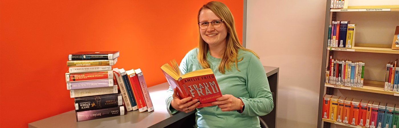 Eine Mitarbeiterin der Bibliothek sitzt an einem Tisch, ein Buch in der Hand haltend