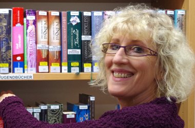 Eine Mitarbeiterin steht an einem Bücherregal und lacht in die Kamera
