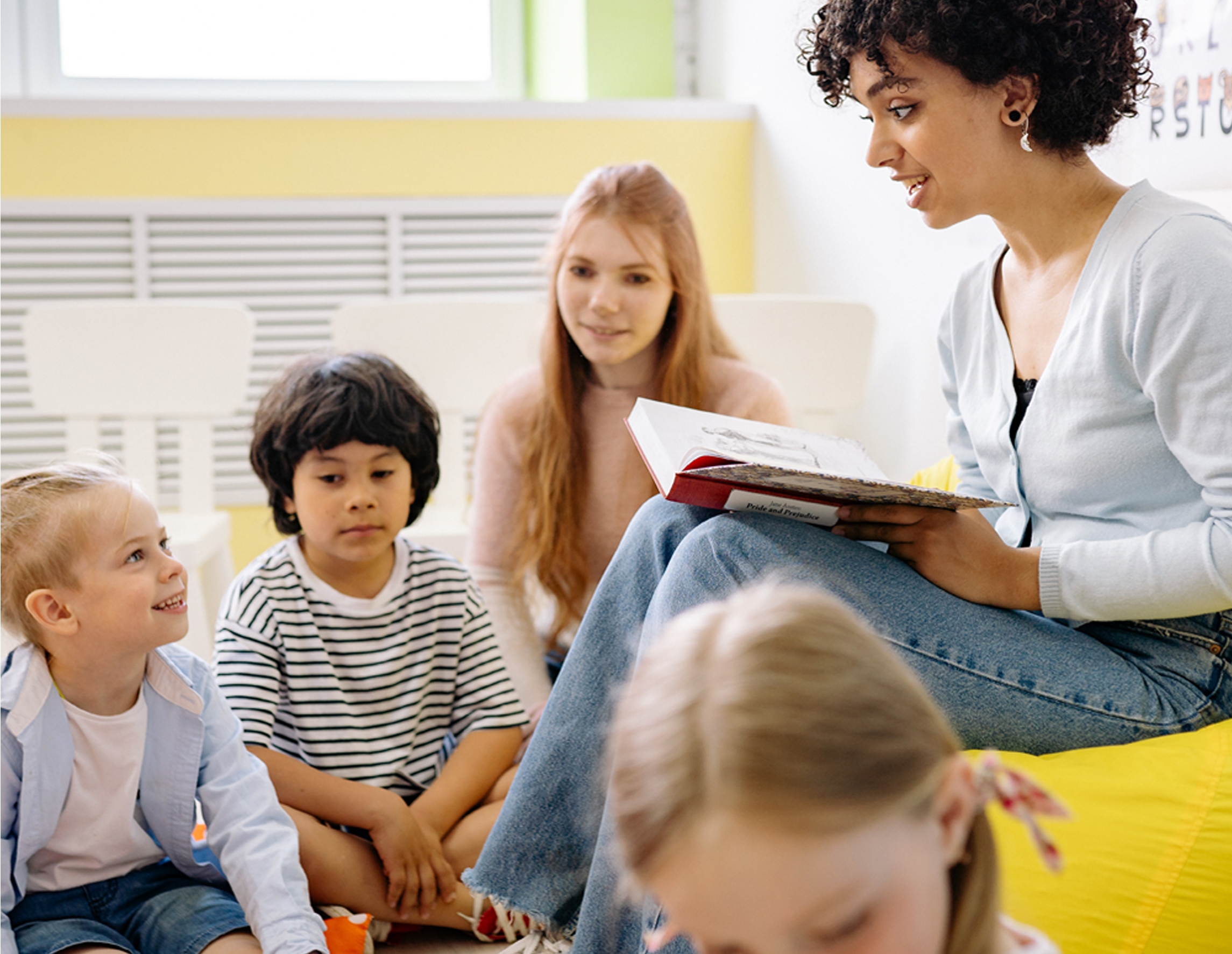Eine junge Frau liest aus einem Buch vor, vor ihr sitzen im Halbkreis Kinder und hören ihr zu