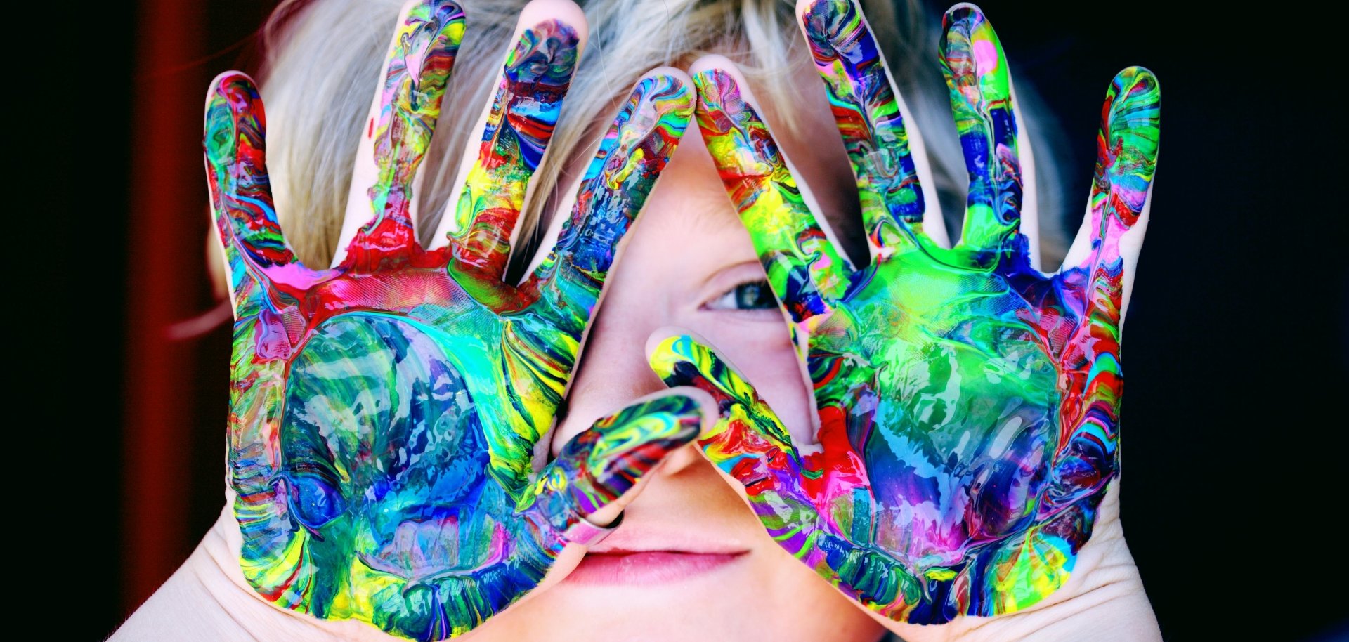 Gezeigt wird ein Kind, das sich mit Fingerfarbe beschmierte Hände vor das Gesicht hält